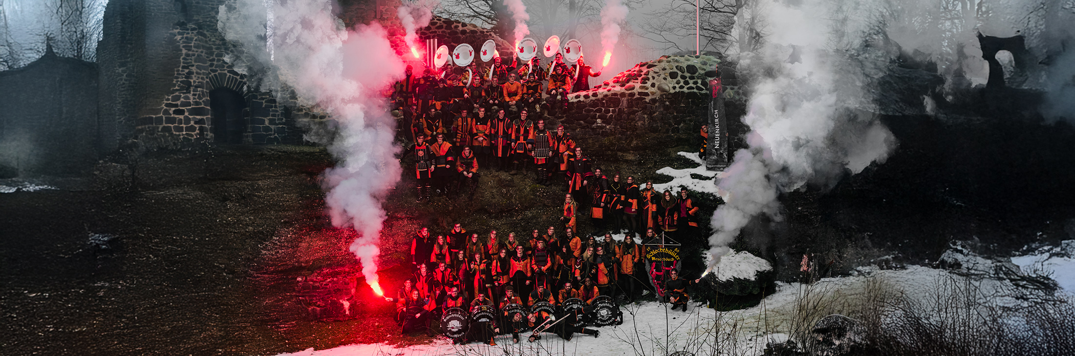 Gesamtbild 2026 der Guggenmusik Schlochthüüler Neuenkirch: Die gesamte Gruppe in rot-schwarzen Kostümen vor einer historischen Steinruine, umgeben von Rauch und roten Fackeln. Das Motto „Mer lönds lah drache“ wird durch eine kraftvolle, düstere Stimmung und drachenhafte Inszenierung visualisiert.