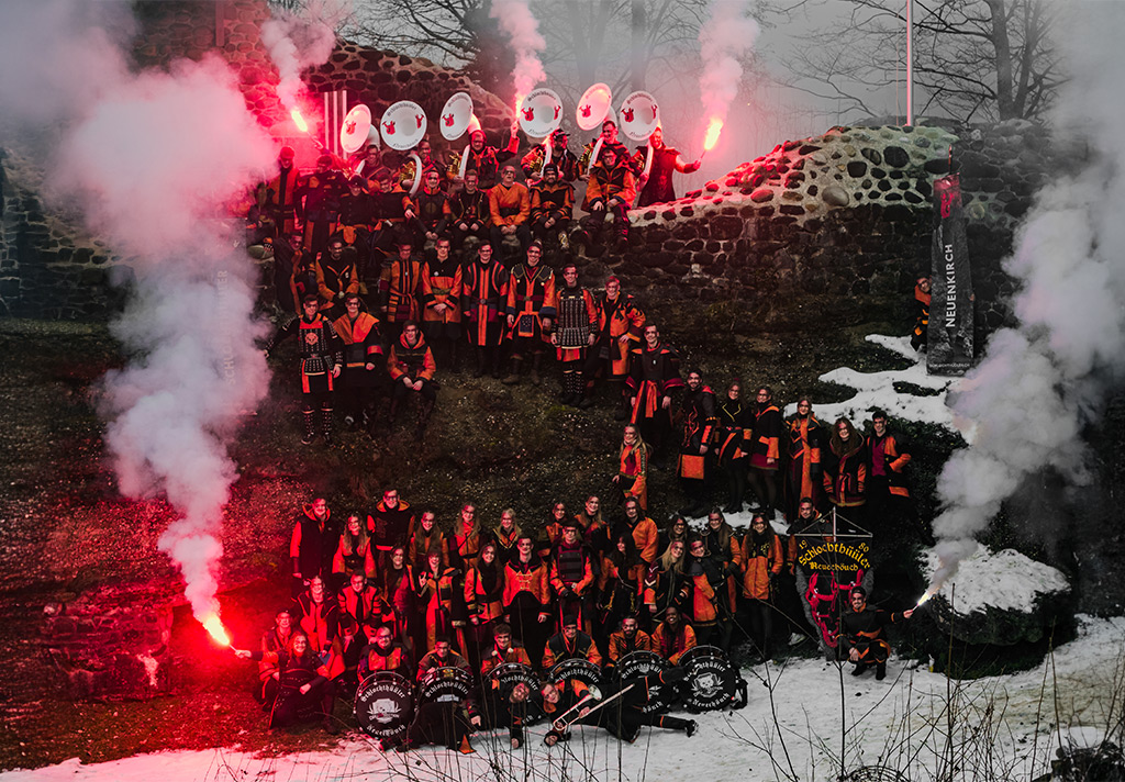 Gesamtbild 2026 der Guggenmusik Schlochthüüler Neuenkirch: Die gesamte Gruppe in rot-schwarzen Kostümen vor einer historischen Steinruine, umgeben von Rauch und roten Fackeln. Das Motto „Mer lönds lah drache“ wird durch eine kraftvolle, düstere Stimmung und drachenhafte Inszenierung visualisiert.